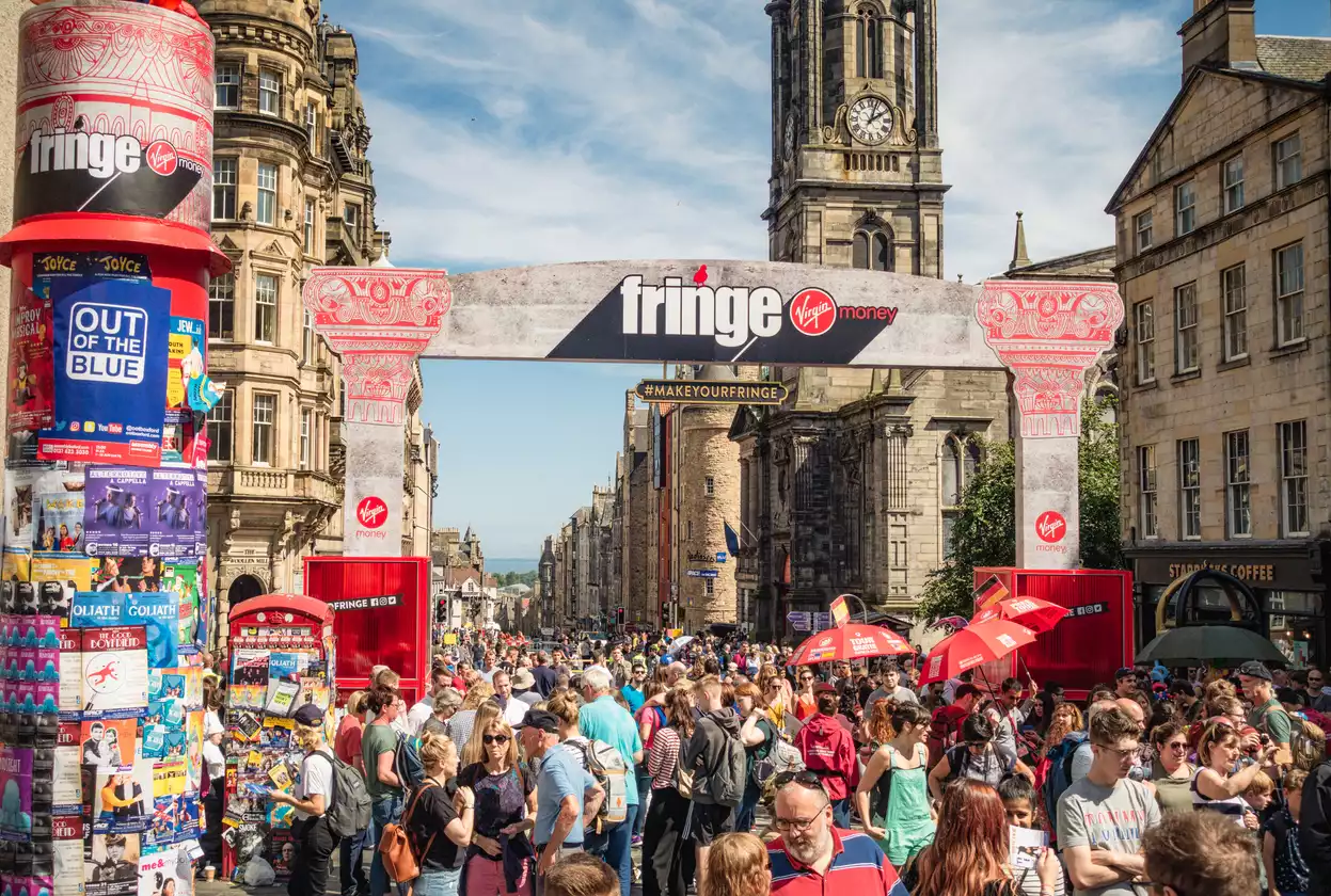 Edinburgh Festival Fringe'in kalabalık bir gününde, tarihi taş binalarla çevrili bir ana cadde boyunca ilerleyen sayısız insan, Virgin Money sponsorluğundaki Fringe yazılı büyük bir kemerin altından geçerken, solda renkli festival posterleriyle kaplı bir stant ve arka planda yükselen bir saat kulesi dikkat çekiyor.