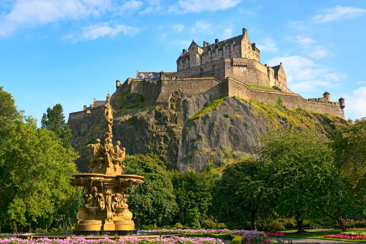 Güneşli bir günde, kayalık bir tepenin üzerinde heybetli bir şekilde yükselen ve eski taş duvarlarıyla dikkat çeken Edinburgh Kalesi'nin, ön plandaki yemyeşil ağaçların, rengarenk çiçek tarhlarının ve zarif bronz bir fıskiyenin bulunduğu bahçe manzarasına tepeden baktığı geniş bir görsel.