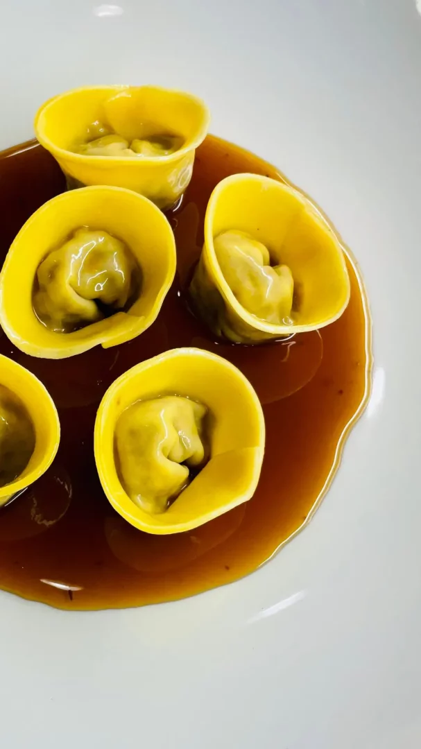 Beyaz bir tabağın üzerinde, iç dolgusu hafif yeşilimsi görünen beş adet parlak sarı renkli, fincan şekilli tortellini veya mantı, zengin ve parlak koyu kahverengi bir sosun içinde özenle servis edilmiş, lezzetli bir yakın çekim sunumu.