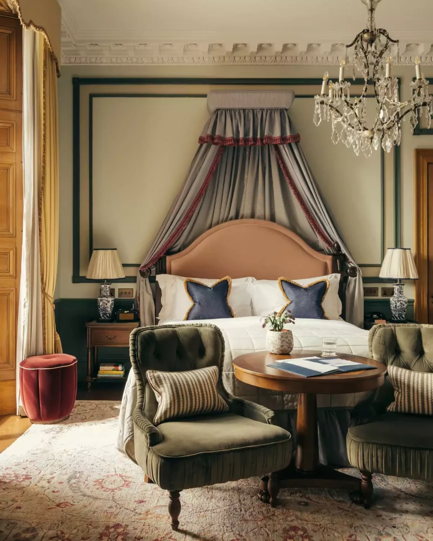 Gleneagles Townhouse'un lüks yatak odasında, somon renkli kavisli başlıklı ve kırmızı şeritli gri kanopili yatak beyaz nevresimlerle kaplı, önünde çizgili yastıklı iki zeytin yeşili kadife koltuk ve ortalarında ahşap yuvarlak bir sehpa bulunuyor.