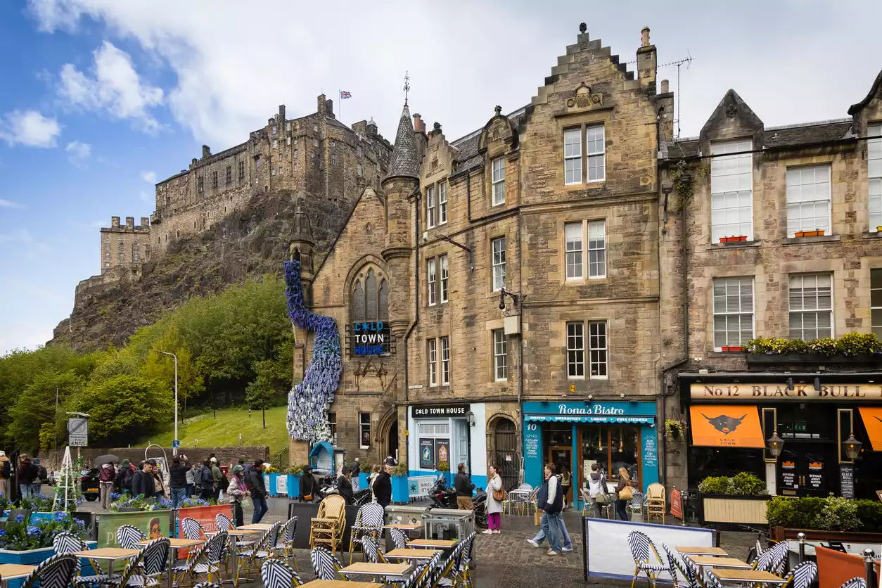 Edinburgh'un tarihi Grassmarket bölgesinde, açık hava yemek masaları, dükkanları olan eski taş binalar ve kısmen bulutlu gökyüzünün altında kayalık bir tepede yer alan Edinburgh Kalesi'nin görüldüğü hareketli bir sokak manzarası.