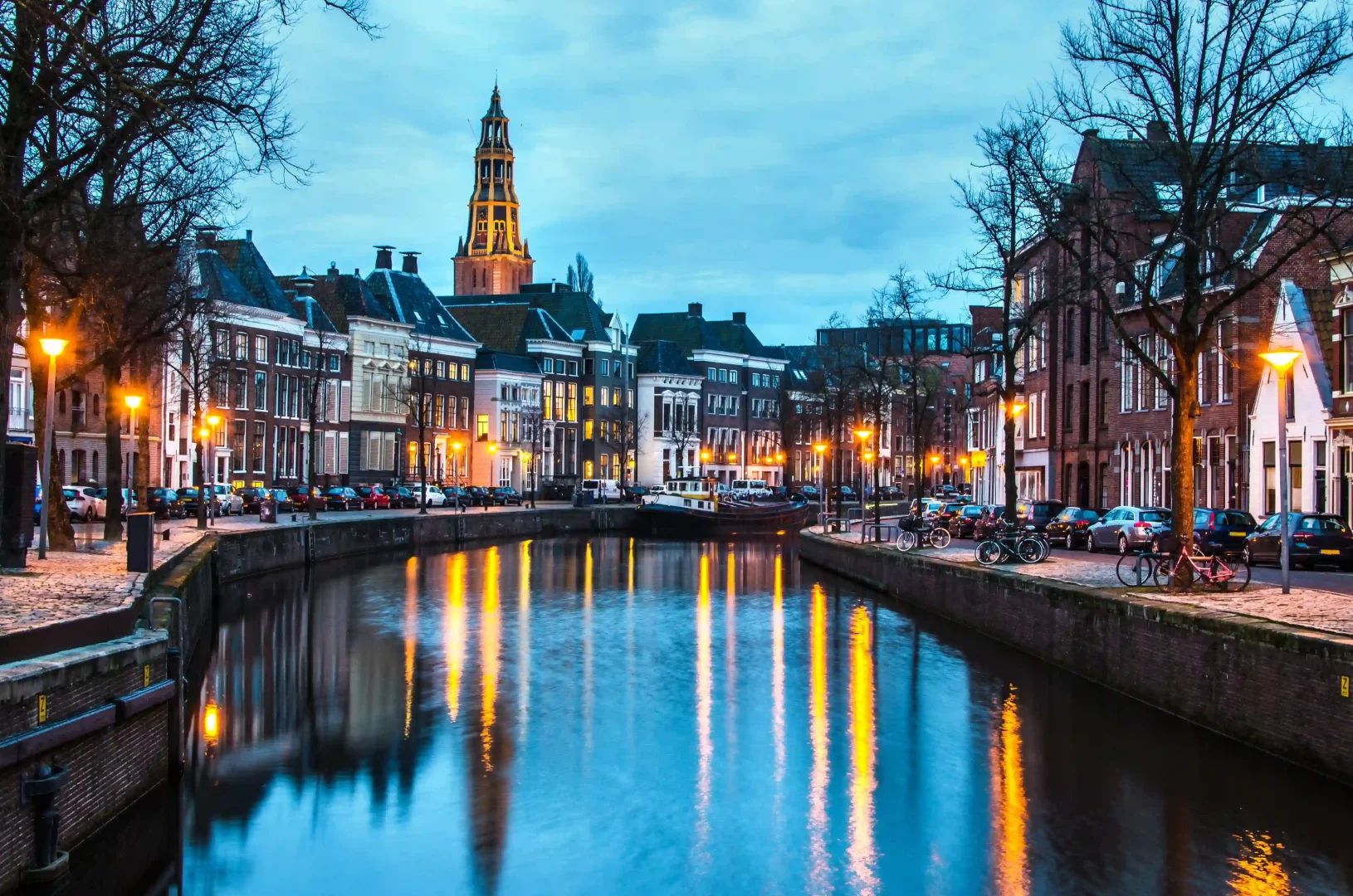 Alacakaranlıkta çekilen bu geniş açılı fotoğrafta, Hollanda'nın Groningen şehrindeki bir kanalın iki yanında sıralanmış geleneksel binaların pencerelerinden ve sokak lambalarından yayılan sıcak ışıklar, durgun suya yansıyarak büyüleyici bir şehir manzarası oluşturuyor ve arka planda kuleli bir kilise göğe yükseliyor.