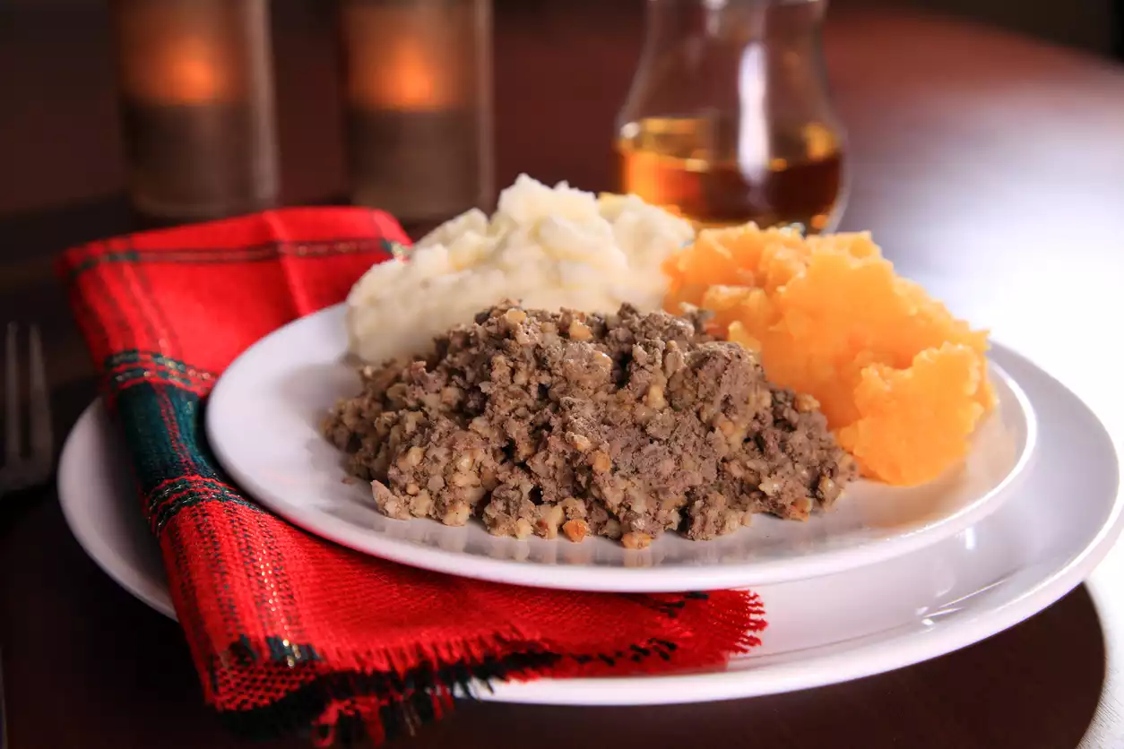 Beyaz bir tabakta, geleneksel İskoç yemeği haggis, püre patates ve şalgam püresi ile servis edilmiş halde duruyor; tabağın altında kırmızı ekose bir peçete, arka planda ise bir viski şişesi ve loş yanan mumlar görülüyor.