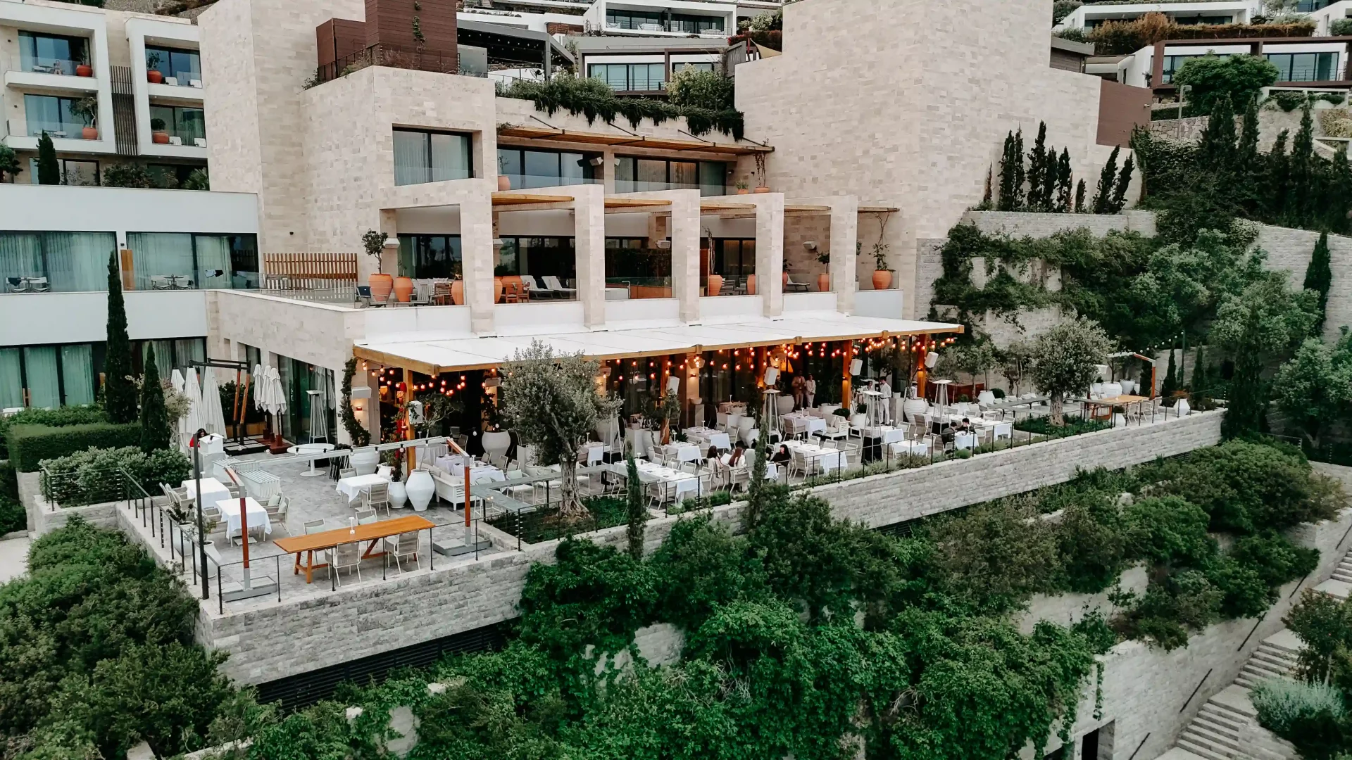 Yemyeşil bir yamaçta konumlanmış modern, taş mimarili bir otel veya tesise ait, ışıklı süs lambalarıyla aydınlatılmış beyaz tenteli geniş bir açık hava teras restoranında, birçok misafirin beyaz örtülü masalarda yemek yediği ve etraftaki zeytin ağaçlarıyla çevrili lüks bir akşam yemeği ortamı.