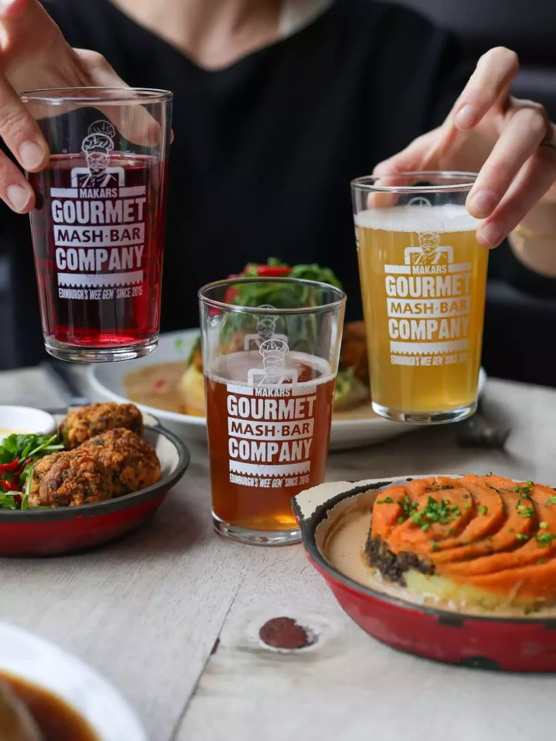 Makars Gourmet Mash Bar logosunu taşıyan iki içecek bardağını havada tutan ellerin göründüğü karede, masada ise haggis topları, püre üzerine tatlı patatesle süslenmiş özel Gourmet Mash yemekleri ve taze salatalar gibi Edinburgh'a özgü zengin lezzetlerin sunulduğu keyifli bir yemek deneyimi yansıtılıyor.