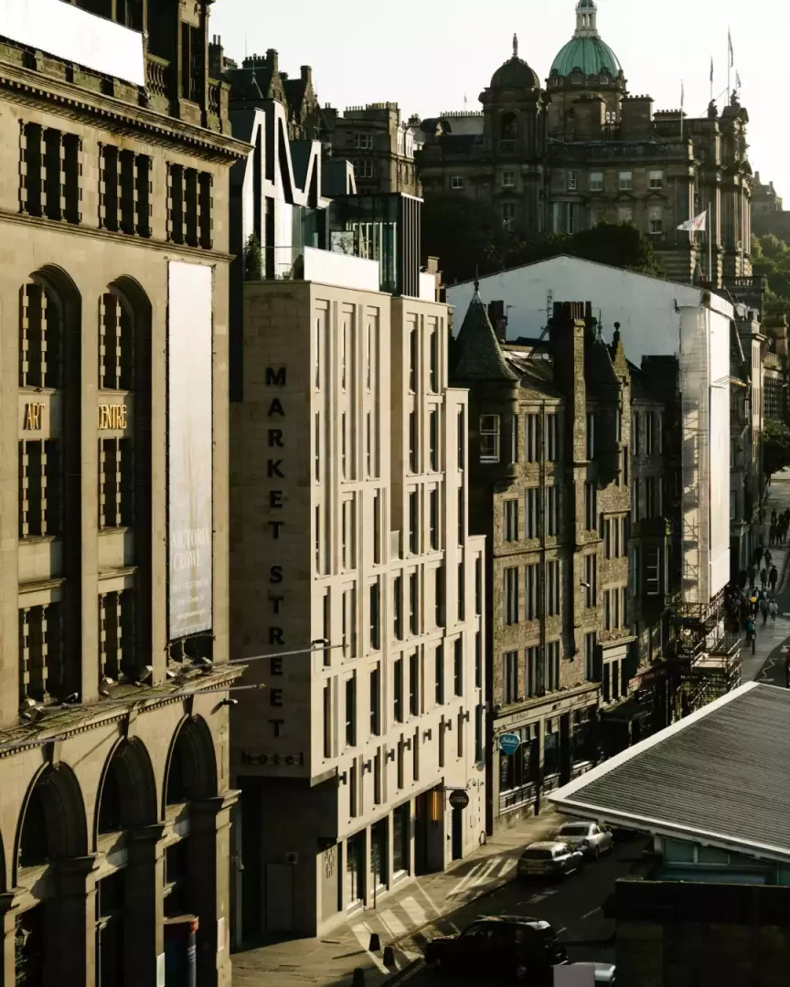 Edinburgh'daki Market Street'te, Market Street Hotel yazısı dikey olarak işlenmiş çağdaş bir bina, kemerli pencereli tarihi Art Centre binası, uzaklarda yeşil kubbeli anıtsal bir yapı ve güneşli caddede hareketli bir şehir manzarasını kuşbakışı gösteren bir fotoğraf.