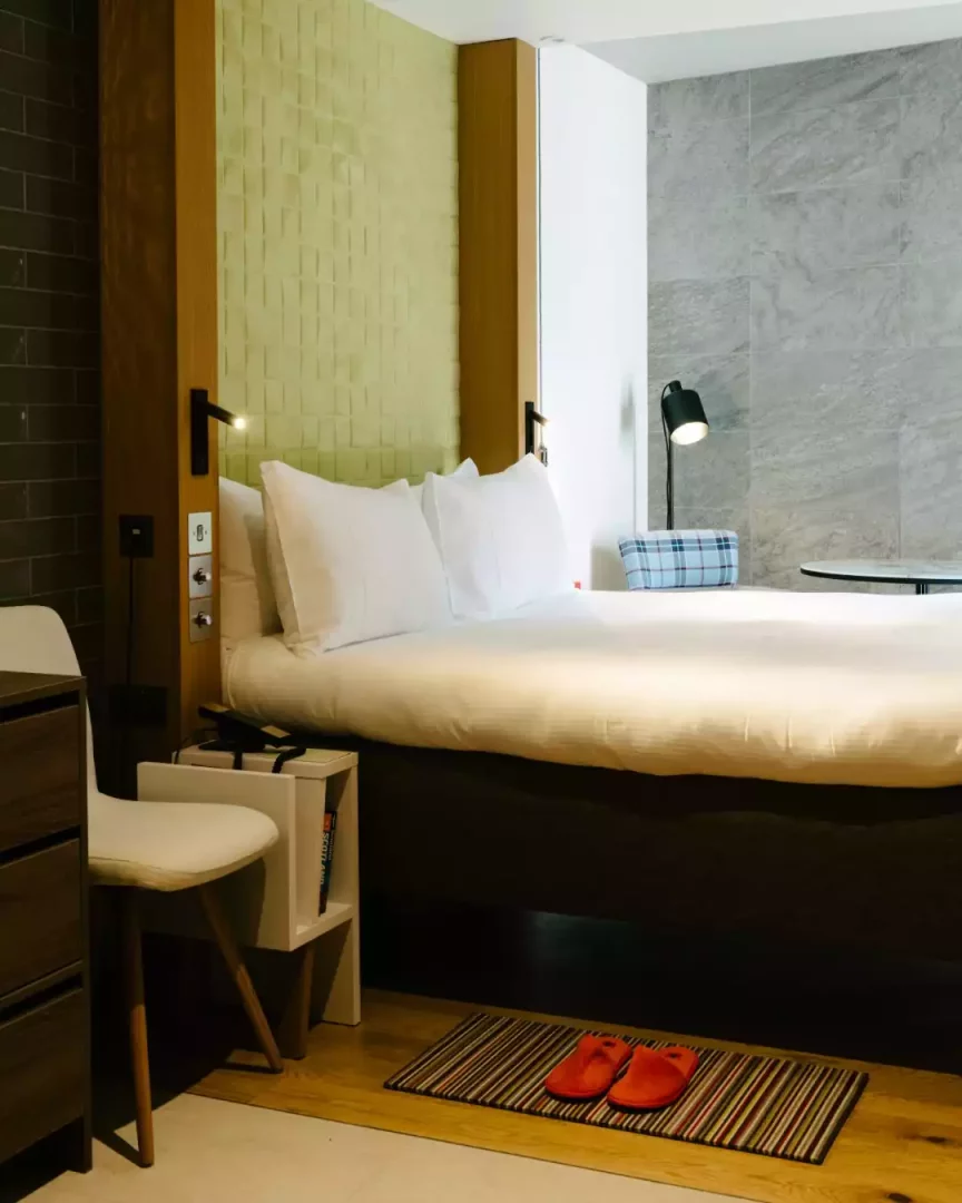 Market Street Hotel'in modern ve konforlu bir otel odasında, yeşil dokulu bir başlık paneli, beyaz çarşaflı geniş bir yatak, yanındaki modern lambalar, ahşap zeminde renkli çizgili paspas üzerinde duran kırmızı terlikler ve sade mobilyalar ile misafirlerini ağırlayan hoş bir atmosfer.