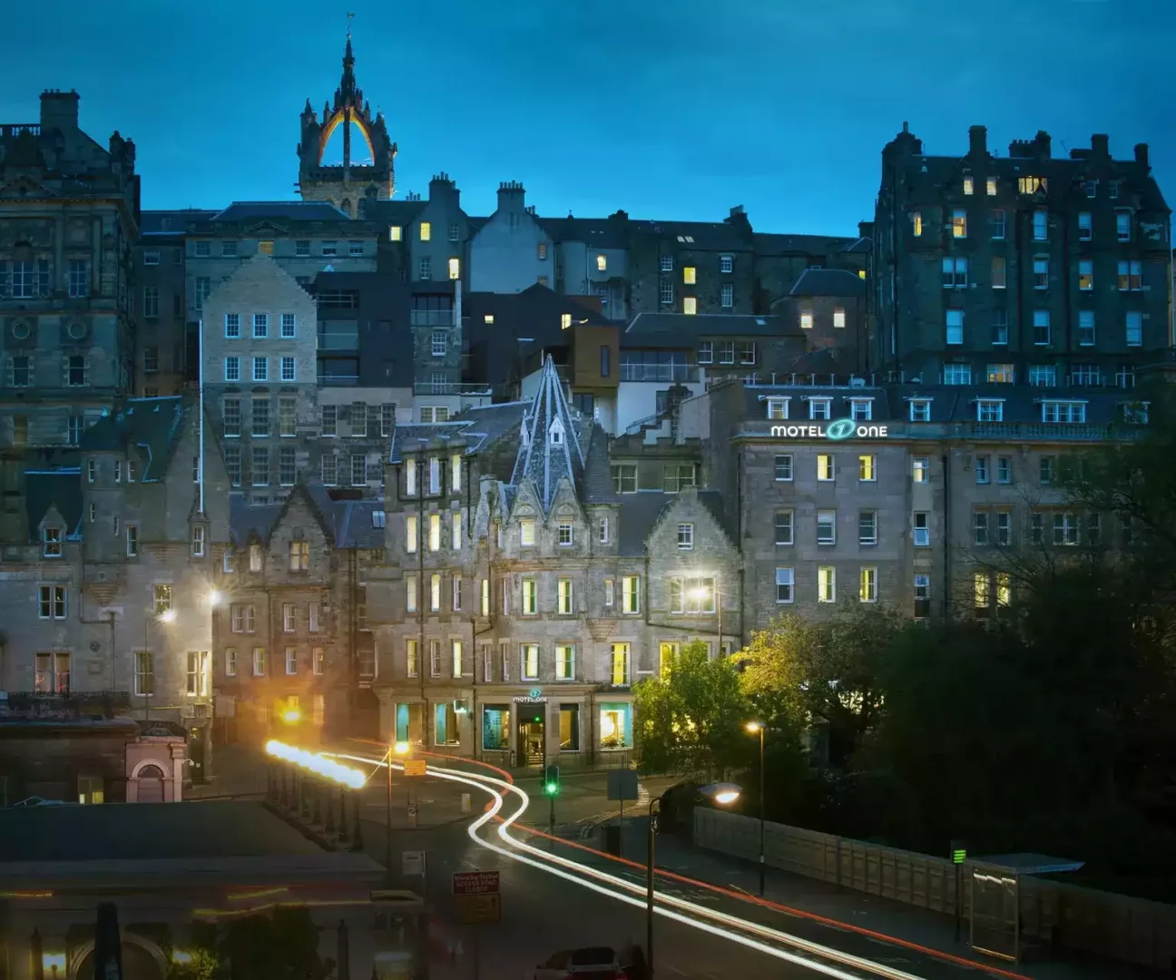 Edinburgh'un tarihi dokusunu yansıtan ve gece ışıklarıyla aydınlanan, hareketli bir cadde üzerindeki binaların arasında yer alan Motel One Edinburgh Royal otelini gösteren uzun pozlamalı bir şehir manzarası.