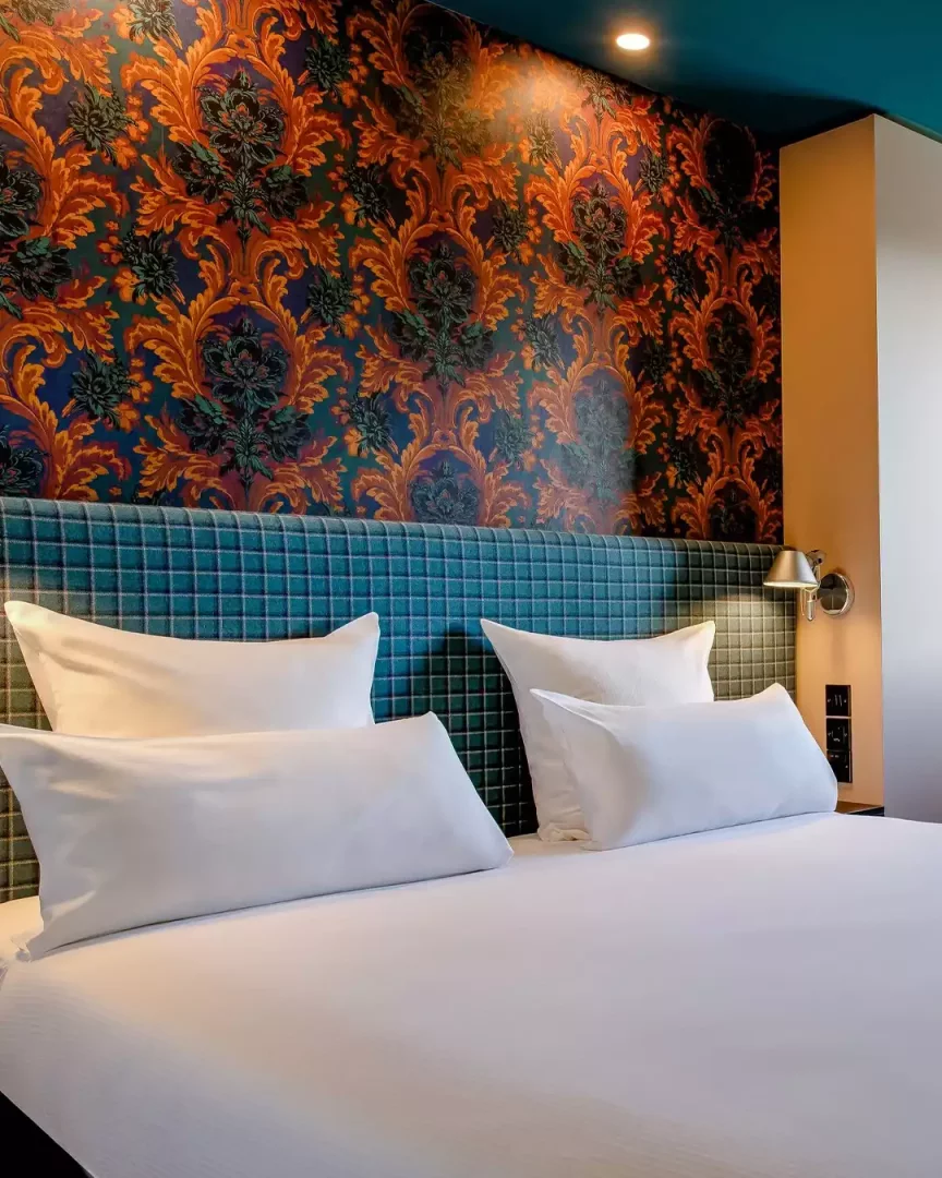 Edinburgh'daki modern bir şehir oteli olan Motel One'da, beyaz nevresimli çift kişilik yatak, kareli mavi yatak başlığı ve üzerinde turuncu, koyu kırmızı, mavi tonlarında geleneksel desenlere sahip duvar kağıdıyla sıcak ve konforlu bir konaklama alanı sunuluyor.