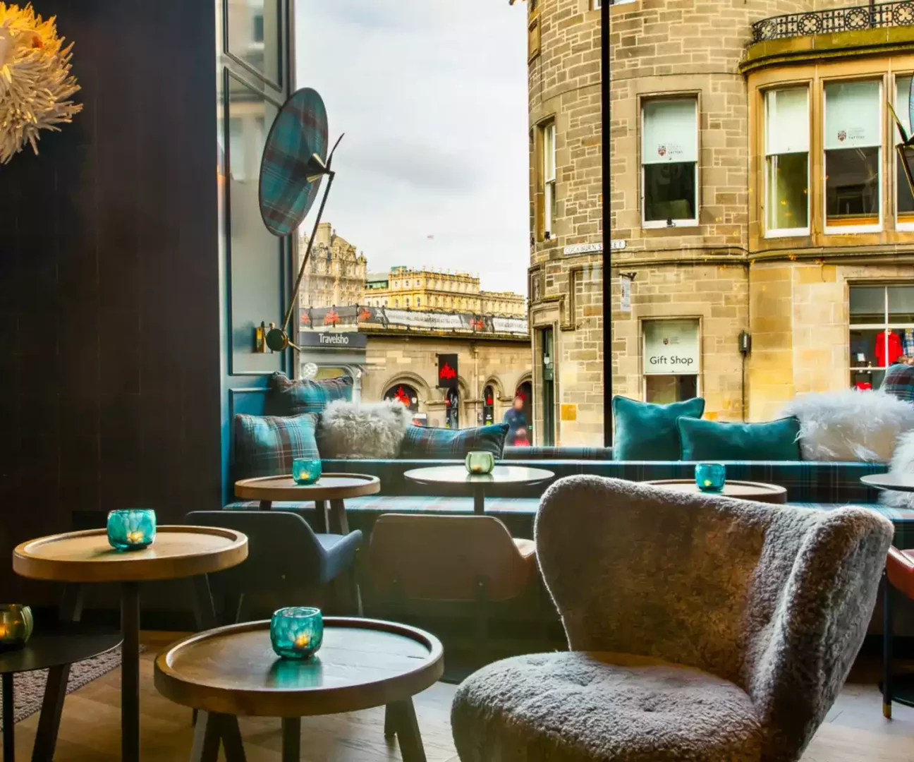 Motel One Edinburgh Royal'ın şık ve sıcak iç mekanında, ekose desenli ve turkuaz minderli uzun banket koltuklar, yeşil cam mumluklarla süslenmiş yuvarlak ahşap sehpalar ve tüylü bir koltuk, dışarıda ise tarihi Edinburgh binaları ve hareketli bir cadde manzarası görülüyor.