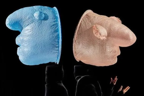 Mummenschanz gösterisinden bir karede, mavi ve pembe tonlarında, abartılı burunlu ve soyut hatlara sahip iki figür, karanlık bir sahnede birbirine dönük bir şekilde duruyor; sağdaki figürün altında bir sanatçının jest yapan elleri görülüyor.