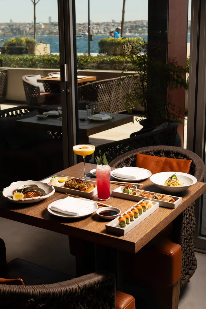Manzaralı bir restoranda, Roka Teishoku deneyimi sunan masada, Japon mutfağının özel lezzetlerinden somon, sushi, gyoza ve ızgara tavuk gibi çeşitli yemekler, ferahlatıcı iki kokteyl ile Boğaz'ın eşsiz manzarası eşliğinde zarif bir şekilde sergileniyor.