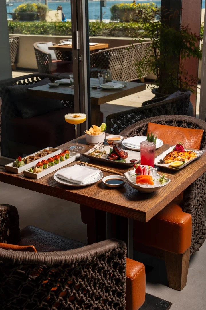 Deniz manzaralı bir terasın önündeki ahşap masada, çeşitli suşi, sashimi, ızgara balık ve et içeren zengin bir Japon mutfağı Teishoku menüsü, renkli kokteyller ve şık servis takımlarıyla çekici bir şekilde sunuluyor.