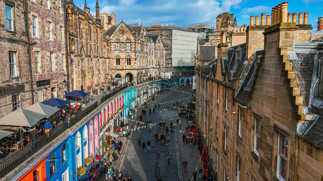 Edinburgh'daki hareketli Royal Mile caddesinde, güneşli bir günde tarihi taş binaların çevrelediği kalabalık bir sokak manzarası; yol boyunca renkli cepheli dükkanlar, açık hava kafeleri ve yürüyüş yapan çok sayıda insan görülüyor.