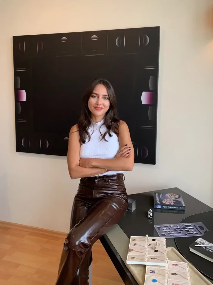 Beyaz kolsuz üst ve parlak kahverengi pantolon giyen, kolları kavuşmuş, gülümseyen bir kadın, soyut bir tablonun önünde, üzerinde 500 EARRINGS adlı kitabın, sergilenen takıların ve bir tasarım şablonunun bulunduğu siyah bir çalışma masasının kenarında oturuyor.