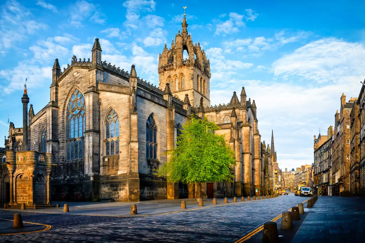 Edinburgh'daki tarihi St Giles Katedrali, Gotik mimarisi ve kendine özgü taç kulesiyle arnavut kaldırımlı bir caddenin solunda, önünde yemyeşil bir ağaç ve sağda eski binalarla aydınlık mavi gökyüzü altında parlıyor.