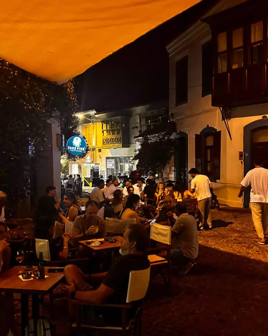 Akşam karanlığında, turuncu bir tente altında, Take Five yazan neon tabelasıyla dikkat çeken bir mekanda insanlar masalarda oturmuş sohbet ederken, çevredeki geleneksel binalar ve Arnavut kaldırımıyla dolu hareketli bir sokak atmosferi.
