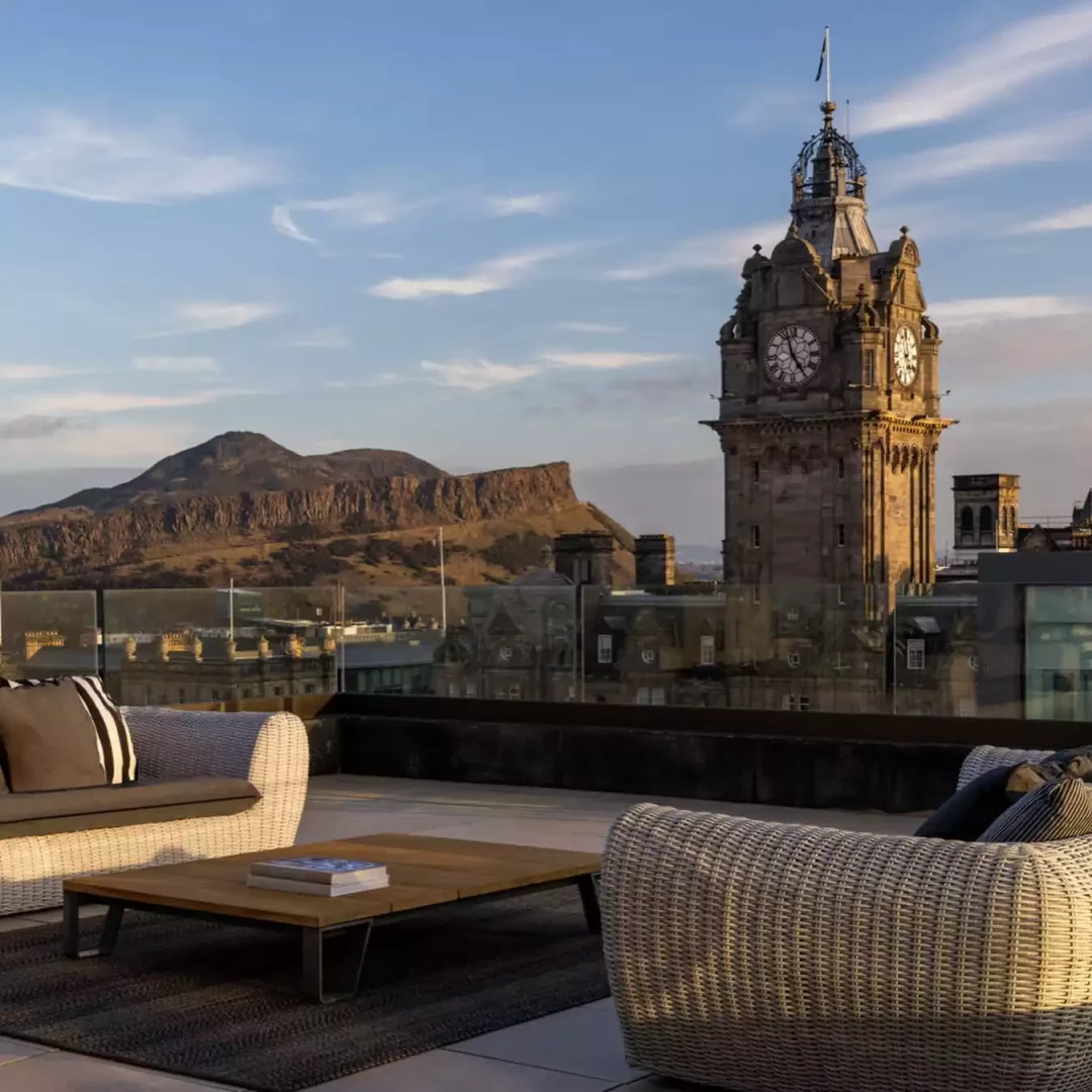 Edinburgh Grand'a ait terasta, ön planda rahat hasır koltuklar ve bir sehpa, arka planda ise Balmoral Saat Kulesi ile Arthur's Seat'in görkemli siluetleriyle Edinburgh şehrinin nefes kesen manzarası güneşli havada göz kamaştırıyor.