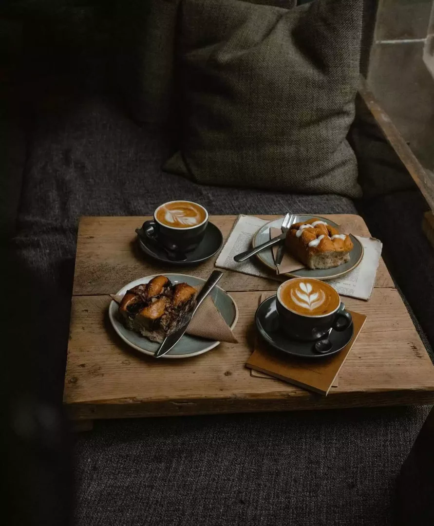 Koyu renkli bir kanepenin önündeki ahşap tepside, özenle yapılmış yaprak desenli latte sanatına sahip iki fincan kahve ile biri tarçınlı diğeri beyaz sırla kaplı iki farklı dilim kekin yer aldığı, sıcak ve samimi bir kahve molası sahnesi.