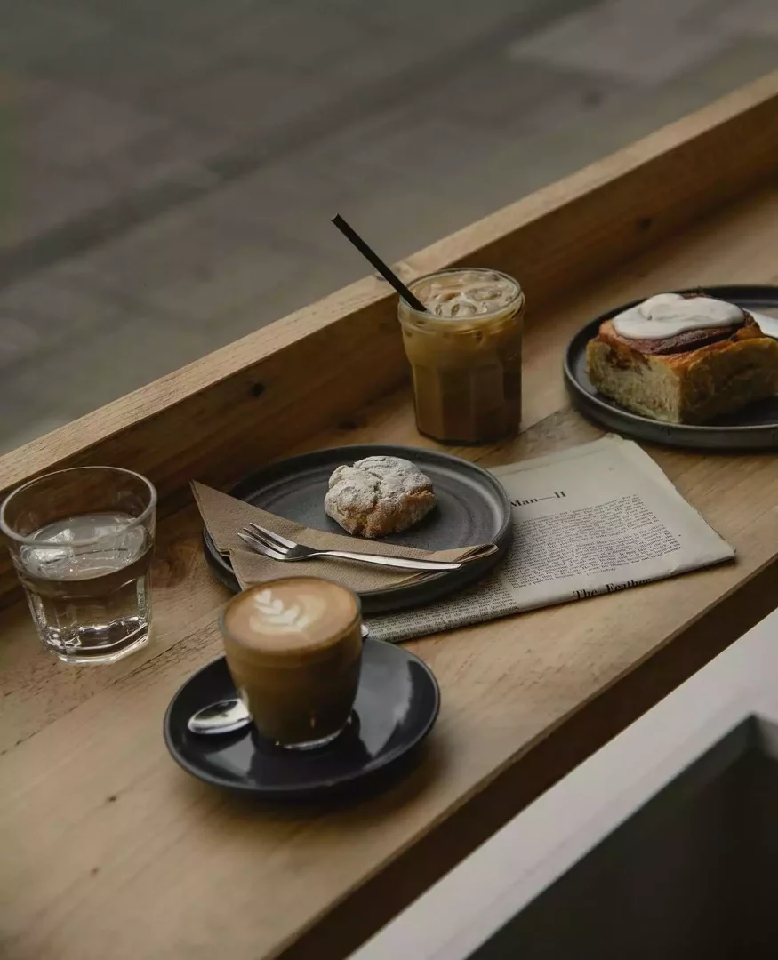 Ahşap bir tezgah üzerinde, süt köpüklü sanatsal bir latte, pipetli buzlu kahve, pudra şekerli bir çörek ve kremalı tarçınlı çörek gibi özenle hazırlanmış çeşitli ikramlar, bir bardak su ve açık bir gazete eşliğinde keyifli bir kafe ortamını gösteriyor.