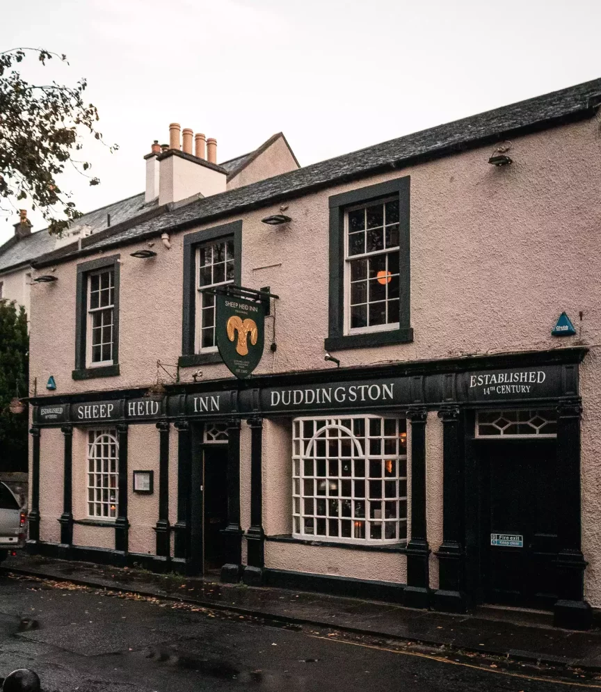 The Sheep Heid Inn Duddingston tabelası ve kuzu başı logosu bulunan, 14. yüzyılda kurulmuş tarihi bir hanın açık pembe cephesi, koyu çerçeveli çok sayıda penceresi ve kemerli geniş vitriniyle yağmurdan ıslanmış kaldırım ve bulutlu bir gökyüzü altında gösteriliyor.