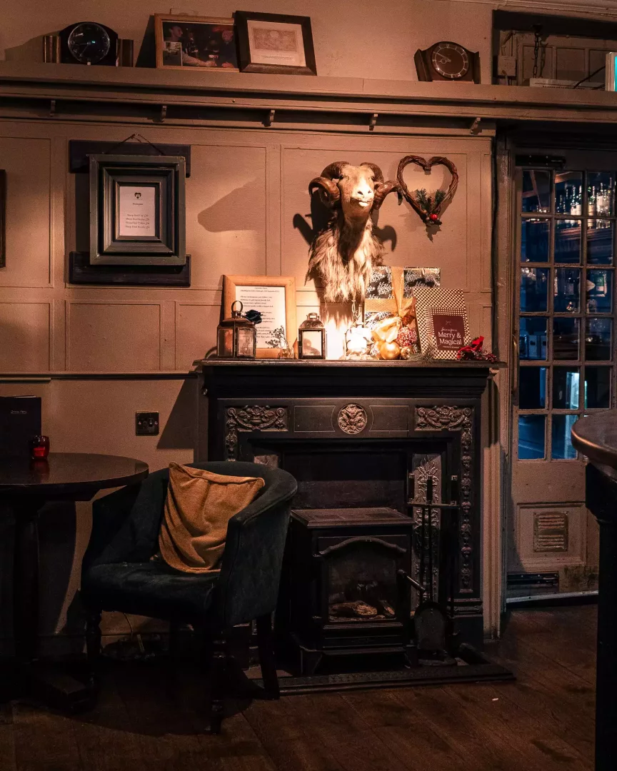 The Sheep Heid Inn'in loş ışıklı geleneksel iç mekanında, görkemli bir şöminenin üzerinde duvara monte edilmiş koç başı, etrafındaki fenerler, hediye paketleri ve kalpli çelenk gibi şenlikli süslemelerle birlikte sıcak bir atmosfer sunarken, şöminenin altında ahşap soba ve solunda rahat bir koltuk yer almaktadır.