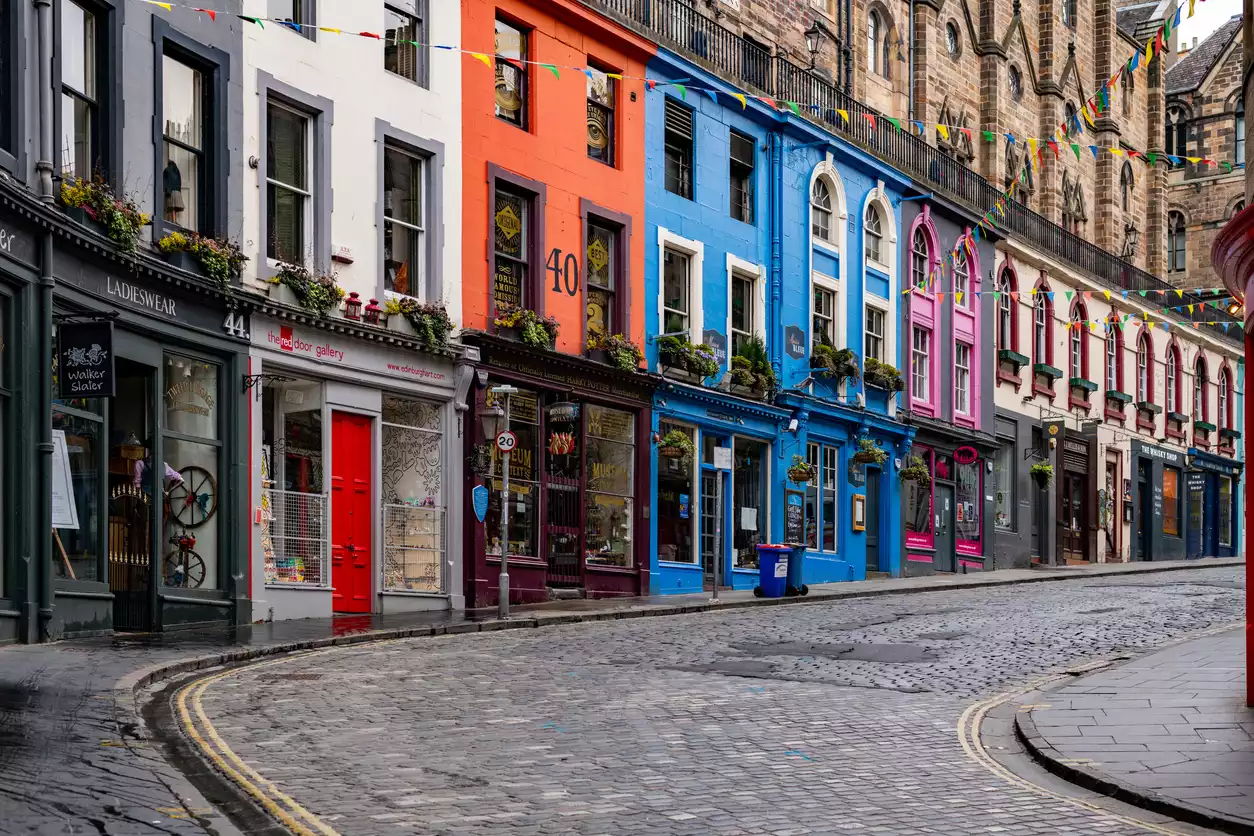 Edinburgh'un ikonik Victoria Caddesi, sıralanmış canlı renkteki dükkan cepheleri, arnavut kaldırımlı kavisli yolu, ıslak yüzeyinde yansıyan ışıkları ve havada asılı rengarenk bayrak süslemeleriyle tarihi ve büyüleyici bir atmosfer sunuyor.