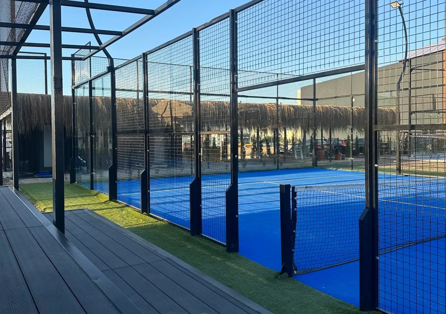 Açık mavi gökyüzünün altında, parlak mavi zeminli, siyah metal ve cam panellerle çevrili bir açık padel kortu, yanlarında yeşil çimler, koyu ahşap bir yürüyüş yolu ve arka planda hasır çatılı dinlenme alanları ile modern ve davetkar bir görünüme sahiptir.