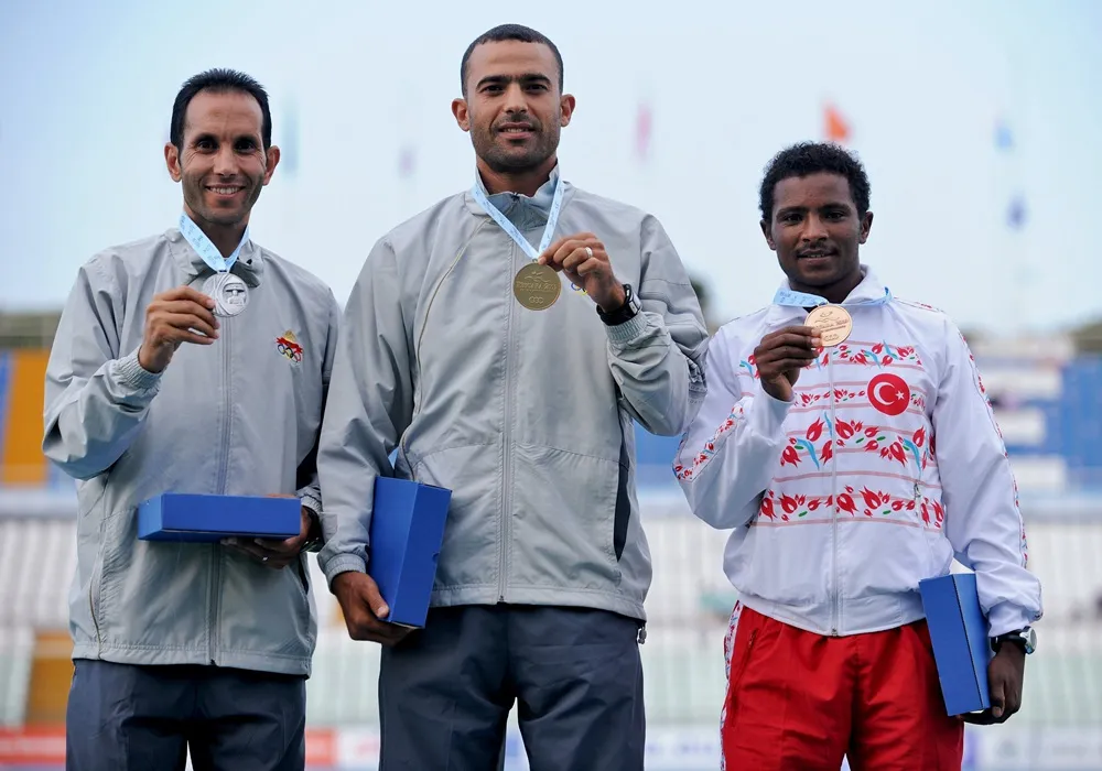 Akdeniz Oyunları'nda, Mersin 2013 logosu taşıyan gümüş, altın ve bronz madalyalarını gururla sergileyen, podyumda kameraya gülümseyerek bakan ve sağdaki sporcunun Türk bayraklı eşofman üstü giydiği üç başarılı atlet, mavi ödül kutularını tutarak zaferlerini kutluyor.