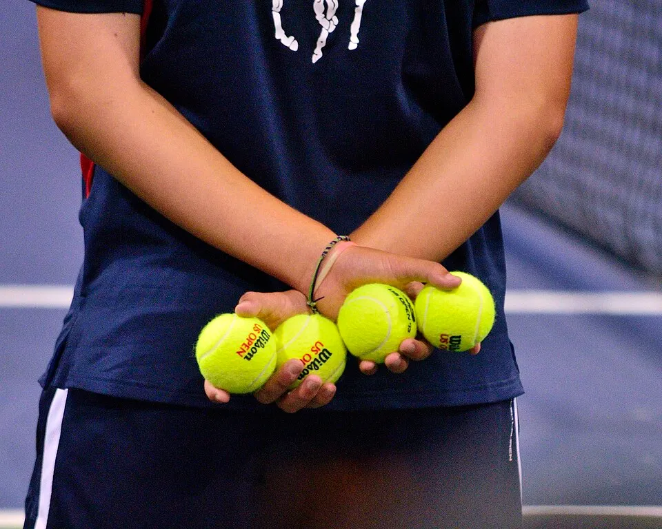 Bir tenis kortunda, koyu renk tişört ve beyaz şeritli koyu renk spor şortu veya eteği giyen bir kişinin ellerinde tuttuğu, üzerinde Wilson US Open logosu bulunan dört adet parlak sarı tenis topu, Amerika Açık turnuvasının canlı atmosferini ve tarihini yansıtıyor.