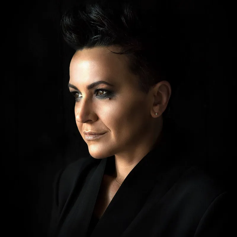 Amira Medunjanin, Sevdalinka ve Balkan müziğini modern yorumlarla birleştiren sahne duruşunu yansıtan bu portresinde, kısa ve üstü kabarık saçları, dikkat çekici göz makyajı ve siyah ceketiyle karanlık bir fonda hafifçe sağa dönük, karizmatik bir ifadeyle yer alıyor.