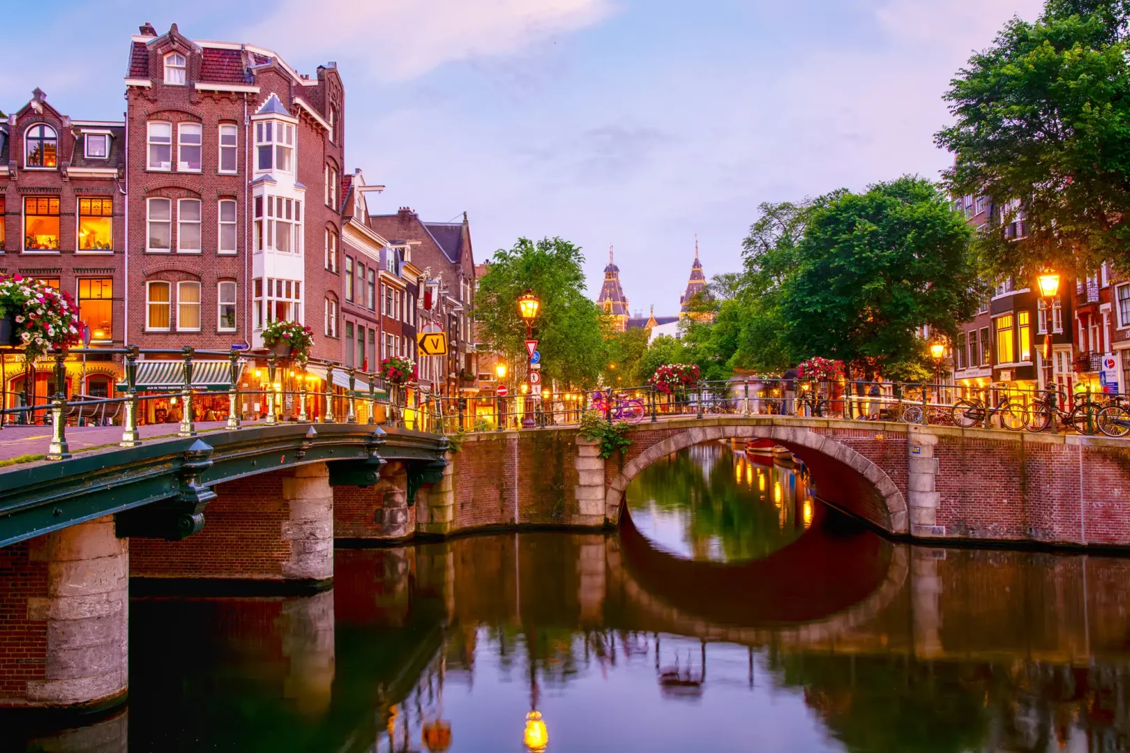 Amsterdam'ın tarihi kanallarından birinde, akşam ışıklarıyla aydınlanmış, taş köprüleri ve çiçeklerle süslü binalarıyla büyüleyici bir görünüm; su üzerindeki yansımalar şehrin atmosferini zenginleştiriyor.