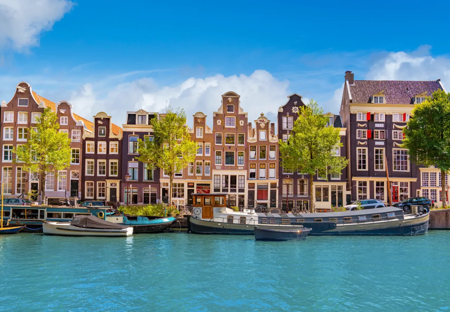 Amsterdam'ın ikonik kanallarından birinin kıyısında, karakteristik gabled çatılara sahip, farklı renklerdeki tarihi kanal evleri, yemyeşil ağaçlar ve üzerinde çeşitli teknelerle birlikte, masmavi berrak bir gökyüzü altında büyüleyici bir şehir görünümü sunuyor.