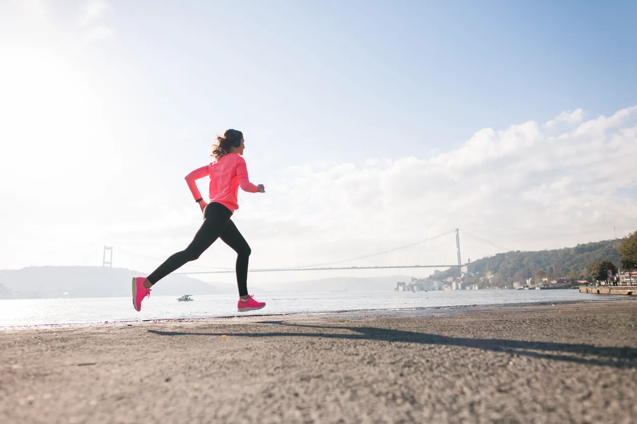 Pembe üstlü, siyah taytlı ve pembe spor ayakkabılı bir kadın, Boğaz Köprüsü manzarası eşliğinde güneşli bir günde sahil yolunda enerjiyle koşarak, Bir Adım Daha Koşusu etkinliğinin temasına uygun sağlıklı yaşamı teşvik ediyor