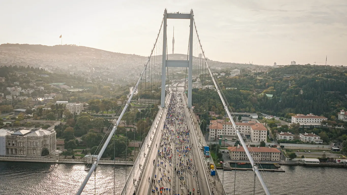 İstanbul Boğazı üzerinde düzenlenen Anadolu Sigorta Bir Adım Daha Koşusu etkinliğinde, Boğaziçi Köprüsü'nü geçen binlerce koşucu, Dolmabahçe Sarayı ve uzaktaki Çamlıca Kulesi'nin de yer aldığı geniş bir şehir manzarası eşliğinde gün ışığında görülüyor.