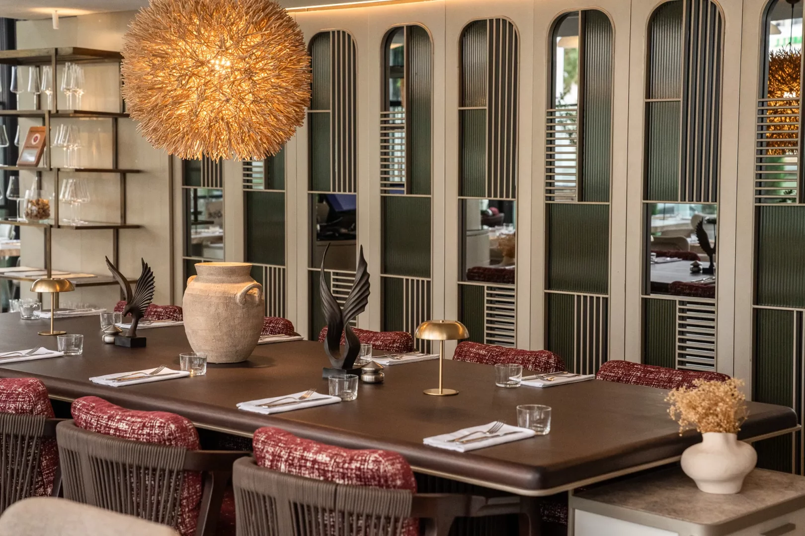 4 Köşe Restaurant'ın modern Akdeniz mutfağı konseptine uygun olarak, sıcak bir ışık yayan küre şekilli büyük avizenin altında konumlanan, koyu renk ahşap masa ve örgü sırtlı, kırmızı desenli minderli sandalyelerle döşenmiş uzun ve davetkar yemek masası, zarif heykeller, rustik bir vazo ve altın rengi lambalarla süslenerek misafirlere keyifli bir gastronomi deneyimi ve uzayan sohbetler için ideal bir ortam sunuyor.