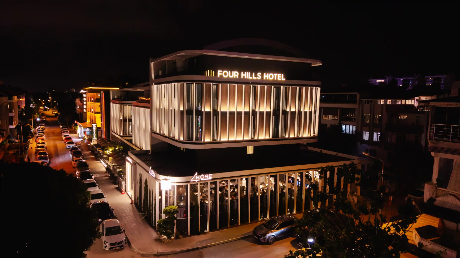 Antalya'da, gecenin karanlığında modern aydınlatmasıyla parlayan, üst katlarında Four Hills Hotel'i barındıran ve zemin katında misafirlerini ağırlayan şık KÖŞE restoranının geniş açılı bir görüntüsü; cadde boyunca park etmiş araçlar ve uzakta seçilen şehir ışıklarıyla tamamlanıyor.