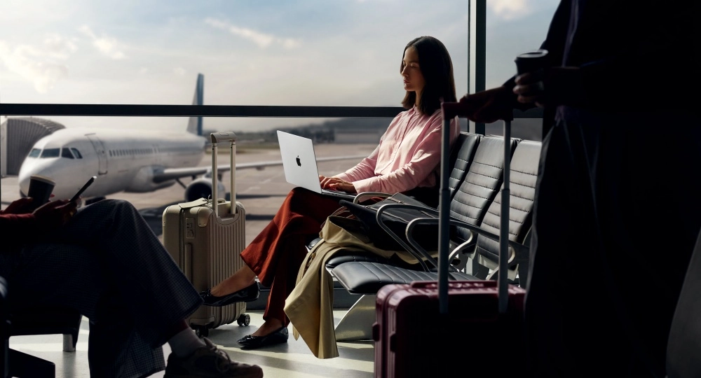 Güneş ışığı alan havaalanı bekleme salonunda, cam pencereden görünen bir uçağın önünde oturan genç bir kadın, gümüş rengi Apple MacBook Pro dizüstü bilgisayarını aktif olarak kullanarak modern, hareketli bir yaşam tarzını sergiliyor.