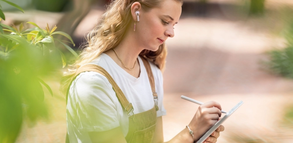 Yeşillikler içinde dışarıda oturan, kablosuz kulaklık takmış, beyaz tişört ve tulum giyen sarı saçlı genç bir öğrenci, elindeki iPad benzeri tablete kalemle dikkatle notlar alarak veya çizim yaparak öğrenci indirimi fırsatlarından faydalanarak pozitif teknoloji kullanımına odaklanıyor.