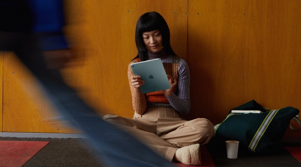 Üniversite ortamında, sıcak tonlarda ahşap bir duvara yaslanmış, çok renkli kazağı ve bej pantolonuyla yere bağdaş kurmuş genç bir öğrenci, elindeki açık mavi Apple iPad'i dikkatle inceliyor; böylece Apple'ın pozitif teknolojiye ve öğrenci indirimlerine olan katkısını vurguluyor.