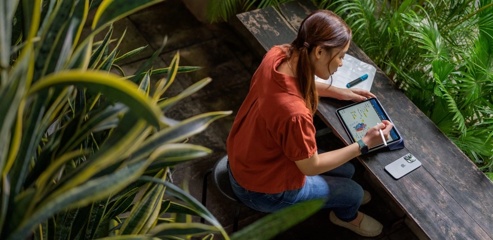 Yeşil bitkilerle çevrili ahşap bir masada oturan genç bir öğrenci, üniversite teknolojileri kapsamında faydalandığı Apple Pencil ile iPad Air'ında biyoloji dersi notları alıp çizim yaparak çalışıyor ve yanında bir iPhone duruyor.
