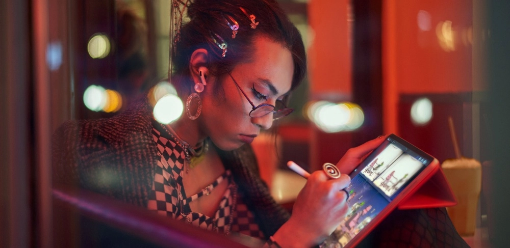 Bir öğrenci, saçındaki renkli tokalar ve gözlüğünün arkasından dikkatle bakarak, kırmızı kılıflı iPad'i ve Apple Pencil ile dijital çizimler yapıyor; arka planda bulanık şehir ışıkları ve bir içecek görülürken, bu an üniversite teknolojileri ve öğrenci indirimi bağlamında pozitif teknolojinin sanatsal kullanımını sergiliyor.