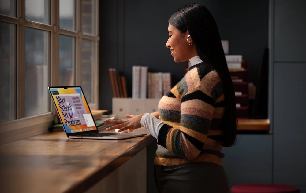 Pencere kenarındaki ahşap bir tezgahta MacBook dizüstü bilgisayarında After School Art Programı'nı inceleyen, AirPods takmış, uzun saçlı genç bir öğrenci, teknolojik araçlarla eğitim ve yaratıcılığa odaklanarak üniversite teknolojilerinin sunduğu fırsatları kullanıyor.