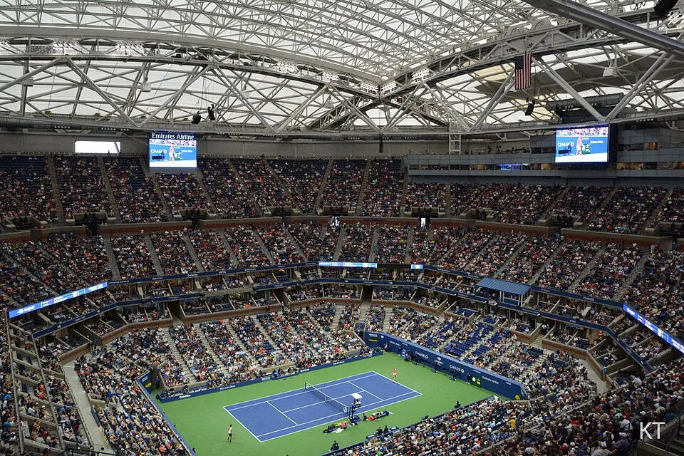Arthur Ashe Stadyumu'nun tavanı kapalı ve karmaşık kafes yapısıyla çevrili devasa iç mekanında, tribünler binlerce tenis severle doluyken, ortadaki parlak mavi kortta oyuncuların heyecanlı bir tenis maçı yaptığı görülüyor; yukarıdaki büyük ekranlarda skorlar ve reklamlar dönüyor.