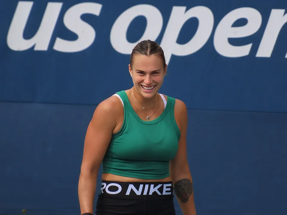 Gülümseyen Aryna Sabalenka, yeşil bir atlet ve siyah şort giymiş, sol kolunda bir kaplan dövmesi varken, arkasında belirgin beyaz harflerle kısmen görünen mavi bir US Open (Amerika Açık) kort duvarının önünde duruyor.