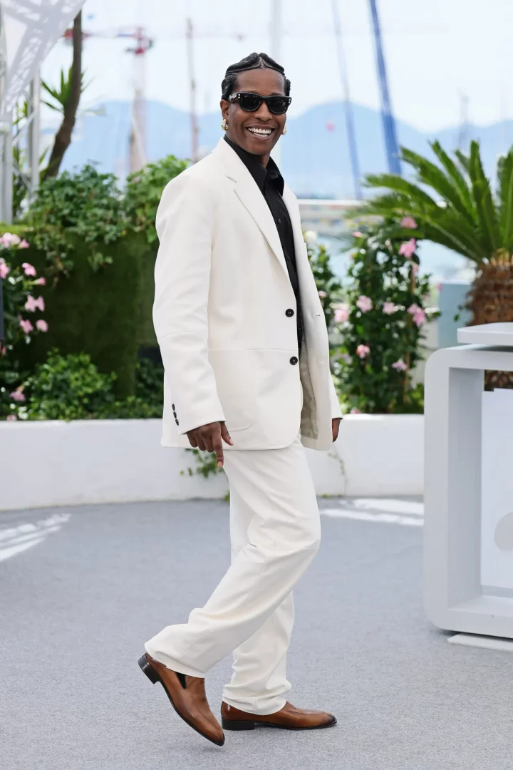 A$AP Rocky, beyaz takımı, siyah gömleği ve güneş gözlükleriyle gülümseyerek objektife bakarken, kahverengi şık ayakkabı modelini sergiliyor ve arka planda yeşillikler ile uzak dağlar dikkat çekiyor.