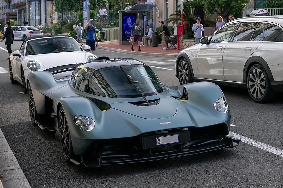 Mat gri-yeşil Aston Martin Valkyrie hiper otomobil, lüks araçlarla dolu yoğun bir şehir caddesinde ön planda, aerodinamik ve keskin hatlarıyla dikkat çekiyor; arkasında beyaz bir Porsche ve sağında bir taksi bulunurken, etraftaki yayalar ve diğer araçlardaki insanlar tarafından hayranlıkla izleniyor.