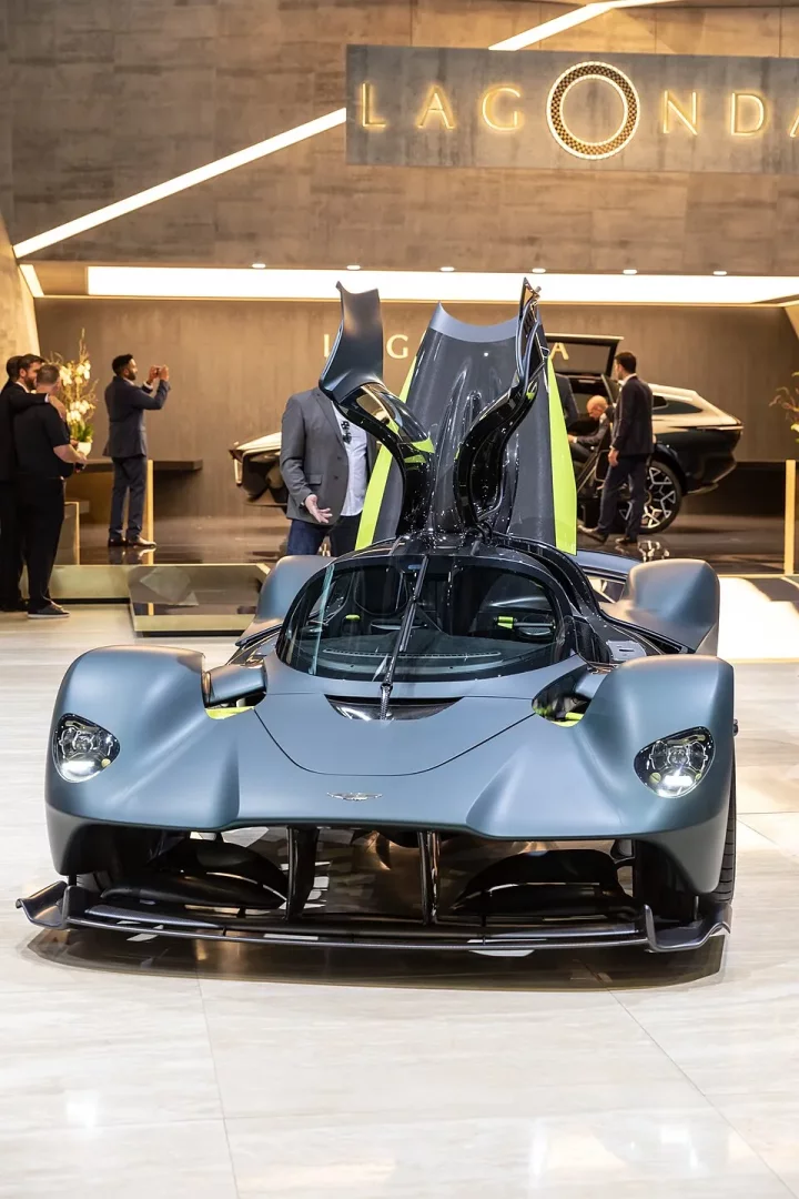 Mat gri rengindeki fütüristik Aston Martin Valkyrie hiper otomobil, kanat gibi açılmış kapıları ve parlak yeşil vurgularla zenginleştirilmiş iç mekanıyla modern bir sergi salonunda, arkada Lagonda tabelası ve aracı hayranlıkla inceleyen veya fotoğraflayan insanlarla dikkat çekici bir şekilde sergileniyor.