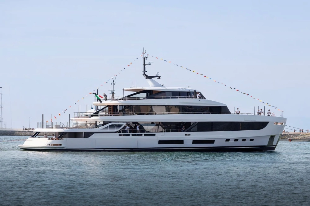 Gökyüzü açık, denizde yüzen beyaz Azimut Grande 44M süper yatının yan profil görünümünde, çok sayıda rengarenk flama ile süslenmiş ve farklı güvertelerinde insanlar bulunan 44 metrelik bu ihtişamlı yatın büyüklüğü ve detayları gözler önüne seriliyor.