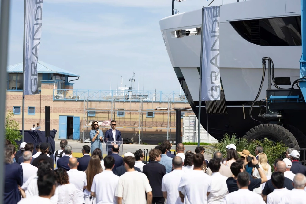 Güneşli bir günde, Azimut Grande 44M super yatın suya indirme töreninde, Grande yazılı bayrakların çevrelediği alanda bir kadın ve bir erkeğin konuştuğu kürsünün önünde toplanan kalabalık ile sağda kısmen görünen 44 metrelik görkemli yeni yatın pruvası.